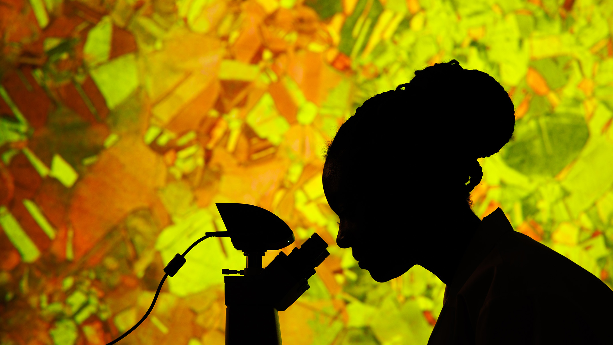 A woman in silhouette looks through a microscope with a backdrop of colored glass.