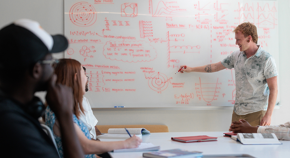 Student writing on a white board with other students sitting nearby looking at the board.