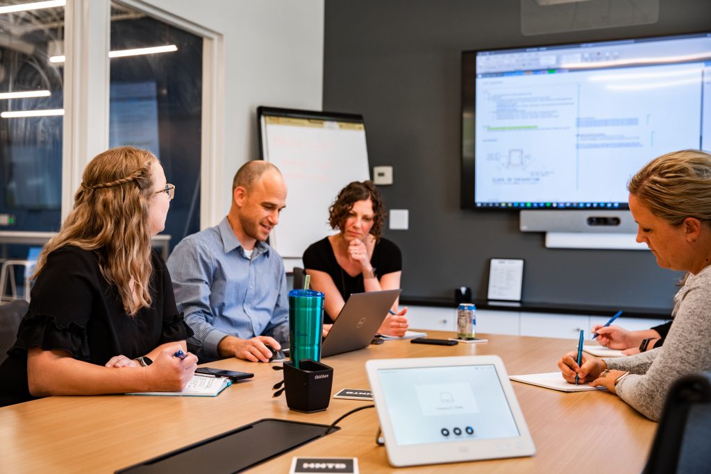 A male student presents from a laptop while two female students looks on, and two more people write on notepads on the other side of the table.