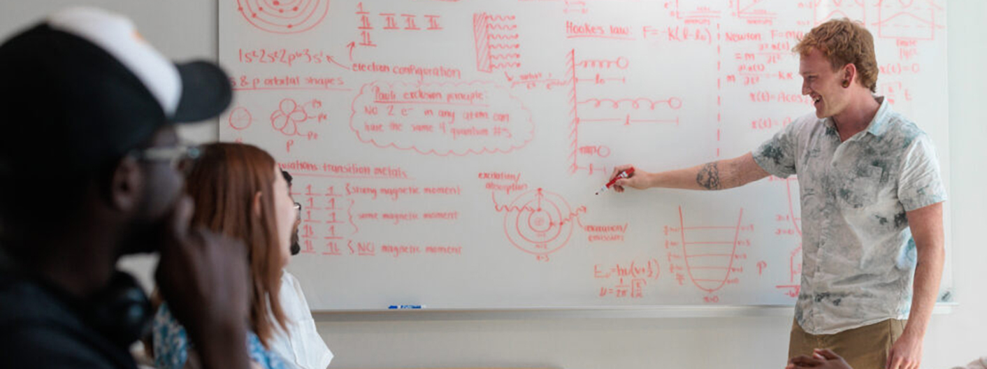 Student instructs classmates at a white board with red writing on it.