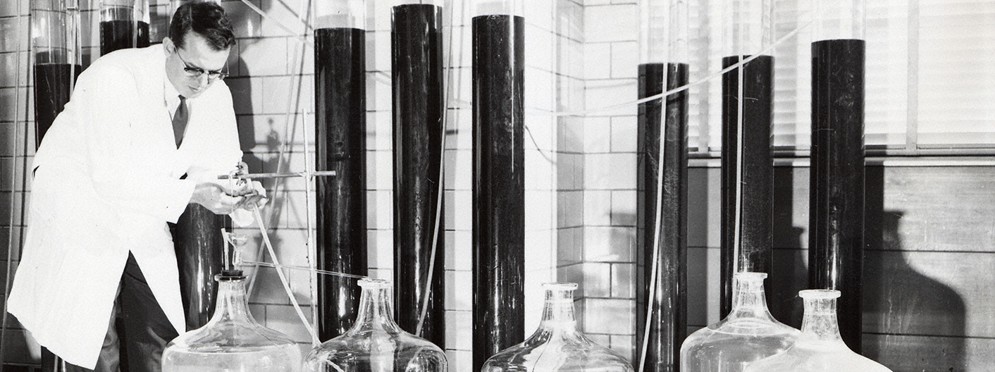 An old, black and white photo depicts a student or professor in a white lab coat working in a metallurgy lab with lots of large glass containers.