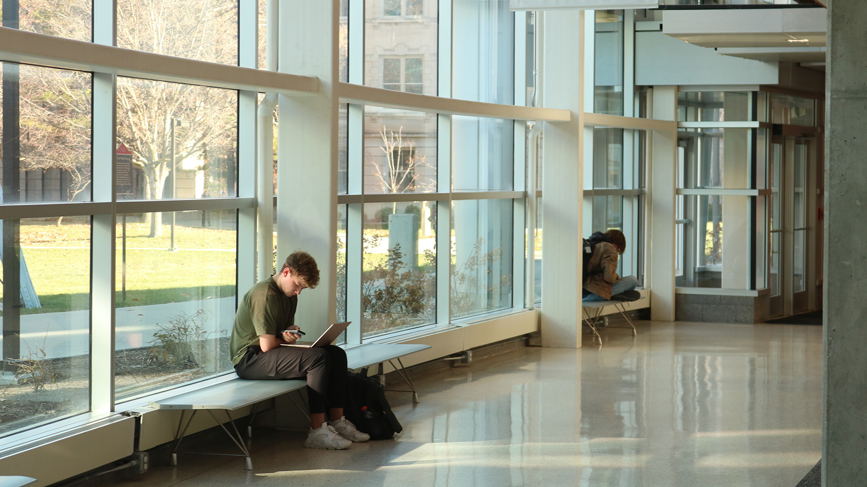 Student sitting and working on a laptop in front of a bank of windows.